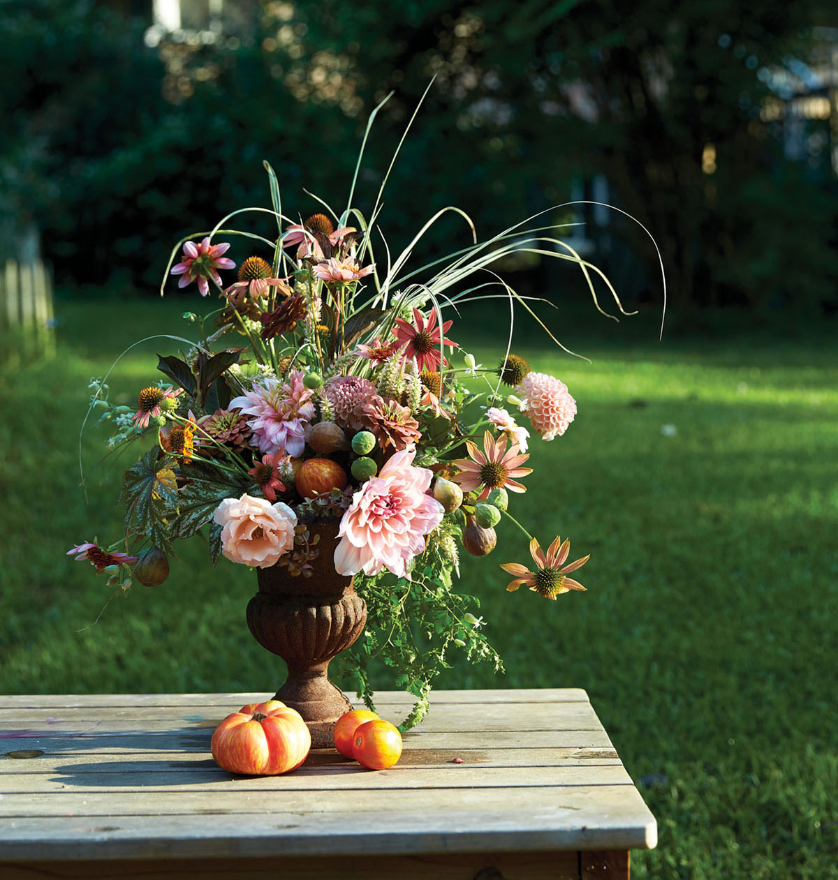 peach, pink, green, and rust-colored summer arrangement by Birmingham, AL floral designer Kappi Naftel of Kap Flowers. The vessel is a rust-colored urn, which sits on a picnic table alongside summer tomatoes, with an expanse of green lawn in the background