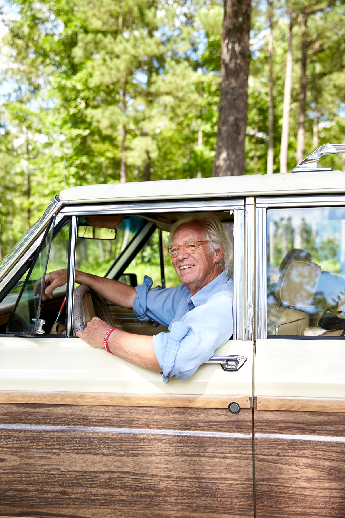 Gates Shaw, wearing a pale blue button up with the sleeves rolled up, smiles from the drivers seat of a vintage wood-panel car